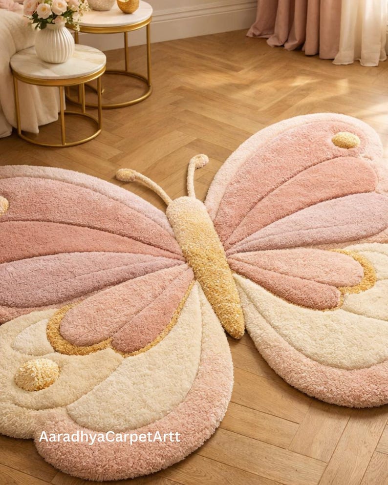 K&ouml;nnte beinhalten: Ein gro&szlig;er, pl&uuml;schiger Schmetterlings-Teppich in Rosa-, Creme- und Gelbt&ouml;nen. Der Teppich hat ein detailliertes Design mit strukturierten Fl&uuml;geln und einem zentralen K&ouml;rper. Der Text "AaradhyaCarpetArtt" ist unten auf dem Teppich zu sehen.