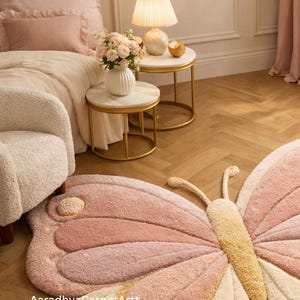 K&ouml;nnte beinhalten: Ein rosa und cremefarbener Teppich in Schmetterlingsform mit strukturiertem Design. Der Teppich liegt auf einem Holzboden, in einem Raum mit Bett, Beistelltischen und einer Lampe. Die Fl&uuml;gel des Teppichs haben verschiedene Rosat&ouml;ne.