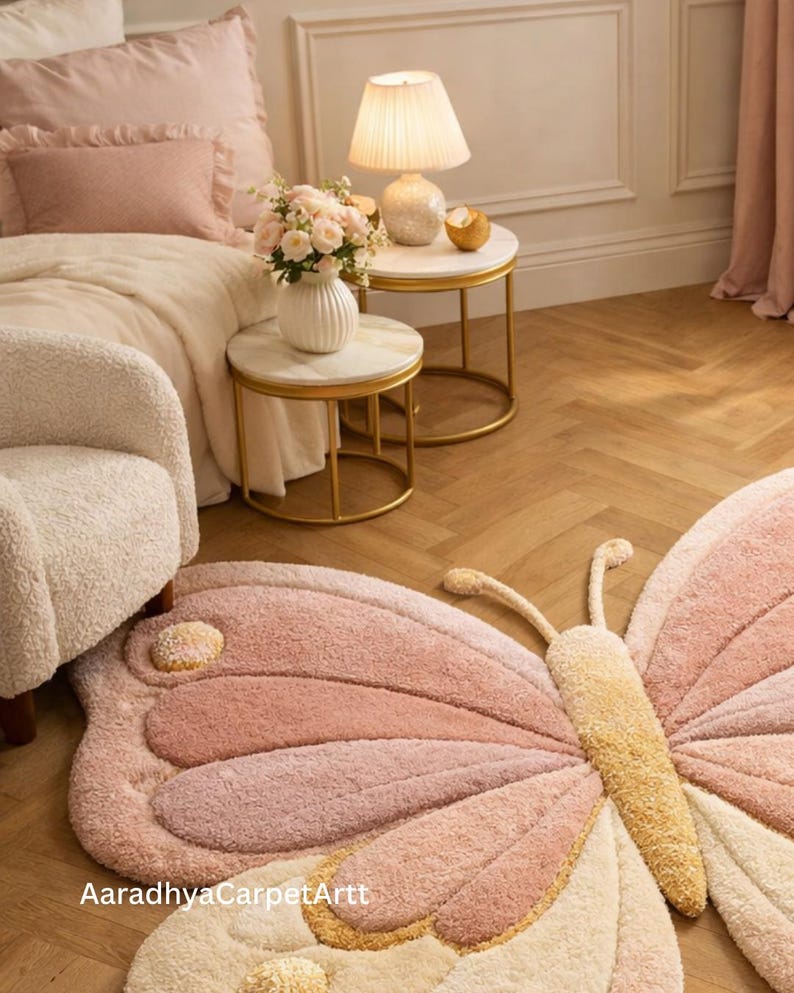 K&ouml;nnte beinhalten: Ein rosa und cremefarbener Teppich in Schmetterlingsform mit strukturiertem Design. Der Teppich liegt auf einem Holzboden, in einem Raum mit Bett, Beistelltischen und einer Lampe. Die Fl&uuml;gel des Teppichs haben verschiedene Rosat&ouml;ne.