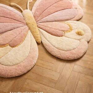 K&ouml;nnte beinhalten: Ein gro&szlig;er, pl&uuml;schiger Schmetterlings-Teppich in Rosa-, Creme- und Goldt&ouml;nen. Der Teppich hat ein detailliertes Design mit strukturierten Fl&uuml;geln und einem zentralen K&ouml;rper. Der Text "AaradhyaCarpetArtt" ist unten sichtbar.