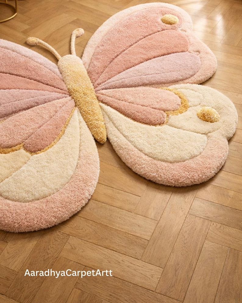 K&ouml;nnte beinhalten: Ein gro&szlig;er, pl&uuml;schiger Schmetterlings-Teppich in Rosa-, Creme- und Goldt&ouml;nen. Der Teppich hat ein detailliertes Design mit strukturierten Fl&uuml;geln und einem zentralen K&ouml;rper. Der Text "AaradhyaCarpetArtt" ist unten sichtbar.