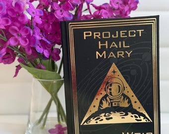 Op de afbeelding: Een hardcover boek getiteld "PROJECT HAIL MARY" van Andy Weir. Het boek heeft een zwarte kaft met gouden letters en een gouden astronautenillustratie. Paarse bloemen in een glazen vaas staan op de achtergrond.