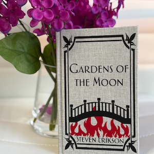 Puede incluir: Un libro de tapa dura titulado "Gardens of the Moon" de Steven Erikson. La portada es de color gris claro con un borde negro y un diseño de puente sobre llamas rojas. Flores moradas en un jarrón de cristal en el fondo.