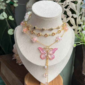 Könnte beinhalten: Ein goldfarbenes Halsketten-Set, das auf einer Schaufensterpuppe präsentiert wird. Das Set besteht aus drei Halsketten: eine mit Sternanhängern, eine mit rosa Edelsteinen und eine mit rosa Sternanhängern. Der Fokus liegt auf einem rosa Schmetterlingsanhänger mit Ketten und Perlen.