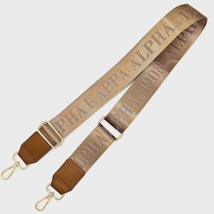 May include: Tan and brown shoulder strap with gold-colored hardware. The strap features the text "ALPHA KAPPA ALPHA 1908" in a repeating pattern. The ends have brown leather accents and gold-colored clasps.