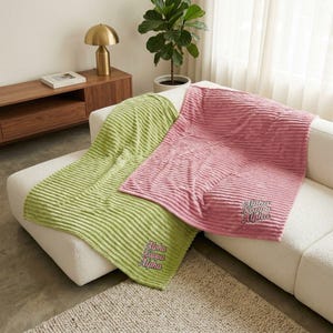 May include: Two corduroy blankets, one light green and one pink, are draped over a white sofa. Each blanket has the embroidered text "Alpha Kappa Alpha" in a script font. The blankets have a ribbed texture.