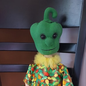 May include: A handmade doll with a green pepper head, yellow tulle collar, and a brown dress with green leaves and yellow and orange fruit designs.