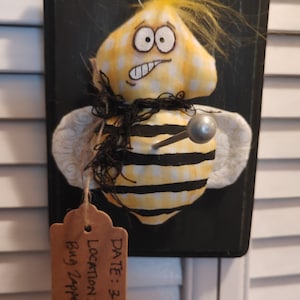 May include: A yellow and black bee with a white face and a black scarf is pinned to a black wooden sign. The sign has a brown tag with the text "DATE: 3-5-22 LOCATION: Bug Zapper".