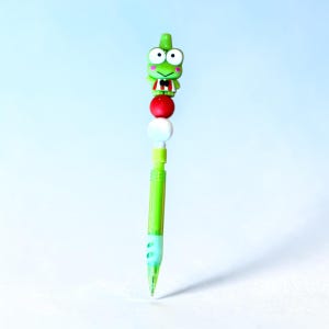 Op de afbeelding: Een groen vulpotlood met een Keroppi-figuurtje als topper. De topper toont een groene kikker met roze wangen die een rood-wit gestreept shirt en een zwarte vlinderdas draagt. Onder het figuurtje bevinden zich een rode en een witte kraal.