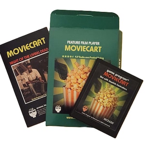 Può includere: Una collezione di prodotti Moviecart, tra cui una scatola verde e due cartucce di gioco nere. La scatola presenta il testo "FEATURE FILM PLAYER MOVIECART" e una grafica di popcorn. Una cartuccia recita "NIGHT OF THE LIVING DEAD". L'altra dice "game program MOVIECART".