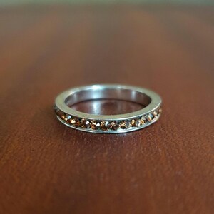 Peut inclure: Une bague en argent ornée d'une rangée de petites pierres précieuses brunes. La bague est posée sur un fond marron, ce qui met en valeur le contraste entre le métal et les pierres. Les pierres sont espacées uniformément.