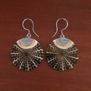 Peut inclure: Une paire de boucles d'oreilles pendantes composées de disques circulaires avec un motif radial marron foncé et crème. Chaque boucle possède un crochet argenté, une section supérieure semi-circulaire perlée et une bande métallique lisse.