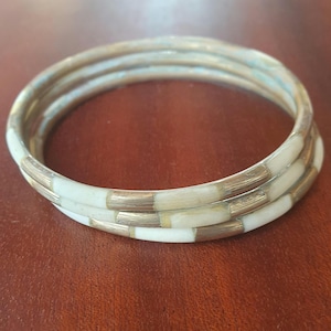 May include: Three stacked bangles with a cream and gold color scheme. The bangles have a rectangular pattern of cream-colored segments separated by gold-colored metal. The bangles are stacked together.