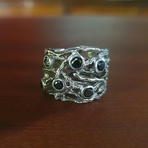 Puede incluir: Un anillo plateado con un diseño texturizado y orgánico. El anillo presenta una serie de piedras preciosas negras engastadas en la intrincada filigrana metálica en forma de ramas. El diseño del anillo recuerda a ramas entrelazadas, creando una estética única y natural.