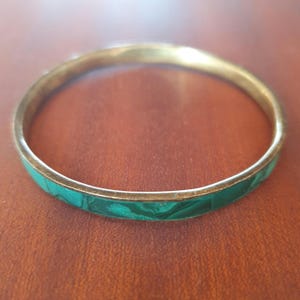 Peut inclure: Un bracelet jonc en laiton avec une incrustation de malachite verte. Le bracelet est de forme circulaire et présente une finition polie. La malachite présente un motif tourbillonnant unique. Un accessoire élégant.