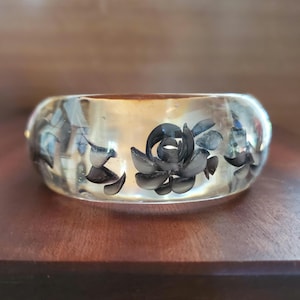 Peut inclure: Un large bracelet jonc en résine transparente incrusté de motifs floraux noirs. La matière translucide laisse apparaître des formes sombres rappelant des roses, disposées en un motif répétitif tout autour de cet accessoire de poignet.