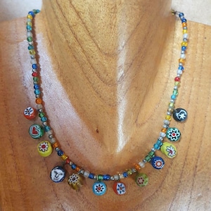 May include: A colorful beaded necklace with small, round, patterned charms. The necklace features a variety of colors, including red, blue, yellow, and green. The charms have floral and face designs. The necklace is displayed on a wooden surface.