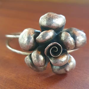 Peut inclure: Bague en métal argenté avec un motif floral détaillé. La bague présente une fleur sculptée avec de multiples pétales et une spirale centrale. Le bracelet est en métal fin et simple. Il s'agit probablement d'un bijou.