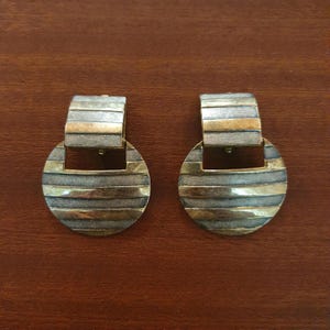 Peut inclure: Une paire de boucles d'oreilles vintage au design géométrique. Chaque boucle d'oreille présente un dessus rectangulaire et un bas circulaire. Les boucles d'oreilles sont argentées et dorées avec des rayures horizontales.