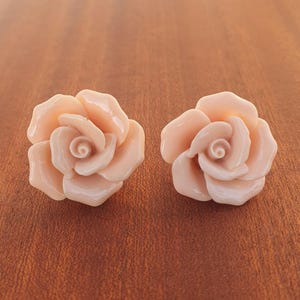 以下が含まれることがあります： 2つの明るいピーチ色のバラの形をした装飾品。各バラには複数の花びらと詳細な中心があります。オブジェクトは茶色の木製の表面に配置されています。バラは滑らかで、おそらく樹脂のような素材でできているようです。