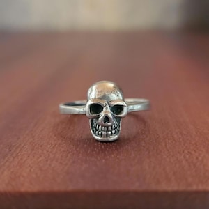 May include: A silver-toned ring featuring a detailed skull centrepiece. The skull has hollow eye sockets, a defined nasal cavity, and visible teeth. The band is thin and smooth, connecting to the sides of the skull, creating a cohesive piece.