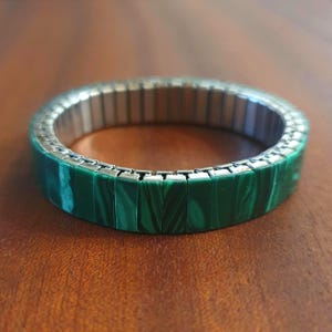 Peut inclure: Un bracelet extensible composé de segments rectangulaires en malachite verte avec des veines sombres naturelles, reliés par une bande métallique argentée flexible. L'accessoire circulaire présente une finition polie sur les pierres.