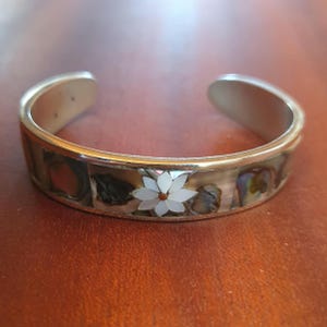 Peut inclure: Bracelet manchette en argent orné d'incrustations de nacre irisée et d'un motif floral blanc. Le bracelet est ouvert. La fleur a un centre blanc et des pétales blancs. Posé sur une surface brune.