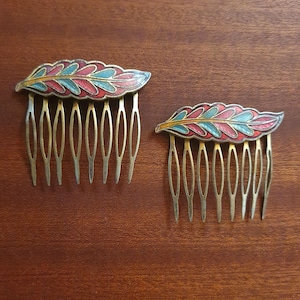 May include: Two hair combs featuring a decorative top shaped like a feather. The feather design is filled with enamel in shades of teal, red, and gold. Each comb has seven metal teeth extending downward from the base of the feather ornament.