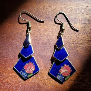 Peut inclure: Une paire de boucles d'oreilles pendantes composées de trois formes géométriques reliées en émail bleu profond. La forme inférieure, la plus grande, présente un motif floral détaillé avec une fleur rouge, une fleur bleue et des feuilles vertes.