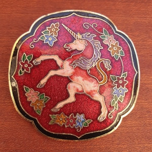 Könnte beinhalten: Dekorative, runde Brosche mit einem Einhorn-Design. Die Brosche hat einen roten Hintergrund mit einem goldenen Rand und einem schwarzen Außenrand. Das Einhorn ist rosa und weiß, mit lila Mähne und Schweif, umgeben von bunten Blumen.