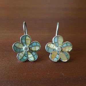 Peut inclure: Une paire de boucles d'oreilles en forme de fleur avec un motif en mosaïque composé de segments bleu clair, vert sauge et jaune pâle. Chaque boucle possède une petite pierre précieuse ronde et transparente au centre et un crochet en métal argenté.