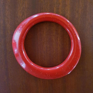 Op de afbeelding: Een cirkelvormige armband in een levendige, glanzende rode tint. Het oppervlak van de armband heeft een gladde, gepolijste afwerking met subtiele, kleine ingebedde spikkels door het materiaal heen, wat zorgt voor een consistente en uniforme uitstraling.