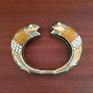 Può includere: Un bracciale rigido in metallo con cerniera a forma di due pesci rivolti l'uno verso l'altro. Il bracciale presenta una finitura oro metallizzato, inserti a griglia quadrata color ambra e piccoli accenti circolari bianchi sul corpo.