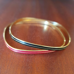 May include: Two gold-tone bangles with enamel accents. One bangle features a red enamel stripe, while the other has a black enamel stripe. Both bangles have a thin gold stripe on either side of the enamel.