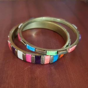 Pode incluir: Dois braceletes circulares de latão com uma série de incrustações retangulares alternadas. As incrustações são coloridas em tons vivos de rosa, roxo, branco, laranja, azul e verde, inseridas entre segmentos metálicos lisos na borda externa.