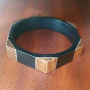May include: A geometric black and wood-tone bangle bracelet. The bracelet has a hexagonal shape with alternating black and wood-colored segments. The wood segments have a natural grain.