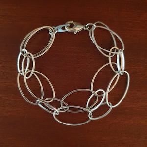 Pode incluir: Uma pulseira de tom prateado construída a partir de uma série de elos metálicos entrelaçados de formato oval. A peça apresenta um acabamento polido e é fixada com um mecanismo de fecho lagosta funcional que une as extremidades da corrente.