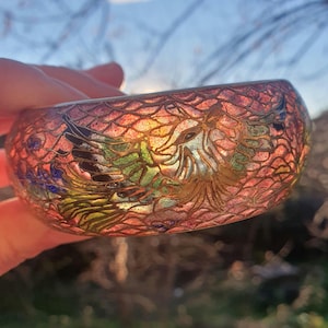 Op de afbeelding: Een kleurrijke, brede armband met een ingewikkeld ontwerp. De armband heeft een gedetailleerd, reliëfpatroon in tinten rood, oranje, groen en goud. Het ontwerp lijkt een mythisch wezen af te beelden. De armband wordt in een hand gehouden.