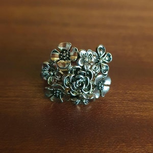 Peut inclure: Une bague en métal argenté composée d'un groupe de fleurs aux formes variées. Le design comprend plusieurs petites fleurs aux pétales et cœurs détaillés, disposées en un bouquet dense et superposé qui recouvre le dessus de l'anneau.