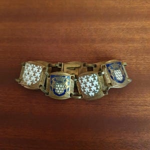 May include: A gold-toned metal link bracelet featuring alternating shield-shaped charms. Two charms display a white field with black ermine spots, while the other two feature a blue shield with a crown, two animals, and the text BRETAGNE.