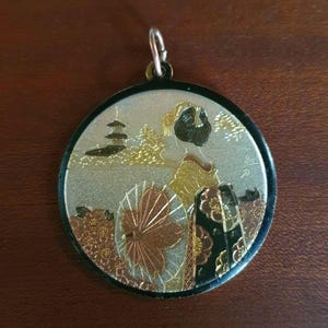 May include: A round metal pendant featuring a detailed engraved scene of a person in traditional attire holding a parasol. The design includes a pagoda, floral elements, and metallic gold, silver, and copper tones with a black border and a silver bail.