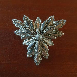 Peut inclure: Une broche en métal argenté présentant un design symétrique en éventail ou en étoile à plusieurs couches. La pièce est composée de nombreux pétales allongés aux bords festonnés rayonnant à partir d'un point central circulaire.