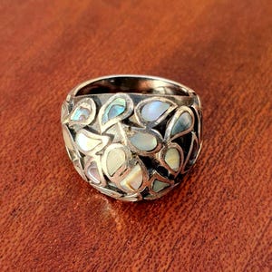 Peut inclure: Une bague en argent au design unique. La bague présente un motif d'incrustations iridescentes en forme de goutte, affichant un mélange de teintes blanches, bleues et vertes. L'anneau est large, et le style général est audacieux et accrocheur.