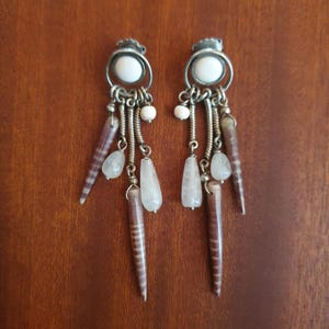 Pode incluir: Um par de brincos pendurados com um design em metal prateado. Cada brinco apresenta uma pedra branca no topo, com elementos pendurados, incluindo pedras brancas e transparentes em forma de gota, pequenas contas brancas e peças alongadas listradas em marrom e branco.