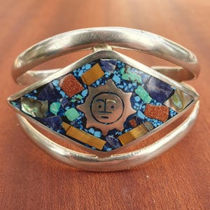 Puede incluir: Pulsera de puño de plata con una pieza central en forma de diamante. La pieza central presenta un diseño de sol de cobre rodeado de un mosaico de piedras azules, turquesas, naranjas y doradas. La pulsera tiene un acabado pulido.