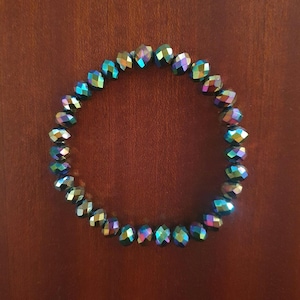 May include: A beaded bracelet with iridescent, faceted beads. The beads display a range of colors, including blues, greens, and purples, creating a shimmering effect. The bracelet is circular and sits on a brown surface.