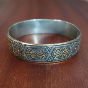 Può includere: Un bracciale rigido circolare in metallo caratterizzato da una finitura scura ossidata con intricati motivi in filigrana color oro. Il design è composto da forme geometriche ripetute, inclusi esagoni e stelle, con dettagliati arabeschi.
