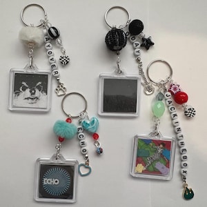 May include: Four square acrylic keychains with metal rings, each featuring unique charms and beads. One displays a dog, one is black, one has a blue optical illusion with the text ECHO, and one shows a person with the text JACK IN THE BOX.