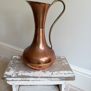May include: A polished copper pitcher with a wide, flared rim and a slender, curved neck. The body is rounded and bulbous, featuring a smooth, reflective metallic surface. A thin, arched brass handle is attached to the side of the neck.