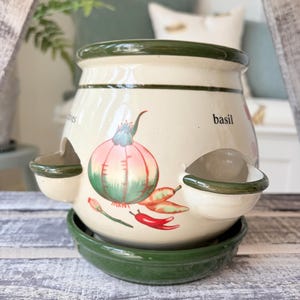 May include: Cream ceramic herb planter with green rim and base. The planter has a colourful illustration of a red onion, chilli peppers, and the word "basil". Two side planters are attached.